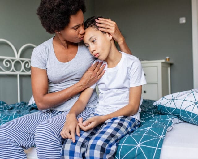 Sad boy is sitting on bed and his mother is hugging and him