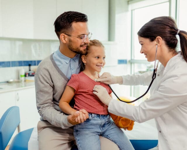 a doctor listening to a child's heart with a stethoscope while the child sits on father's lap in a pediatric urgent care setting