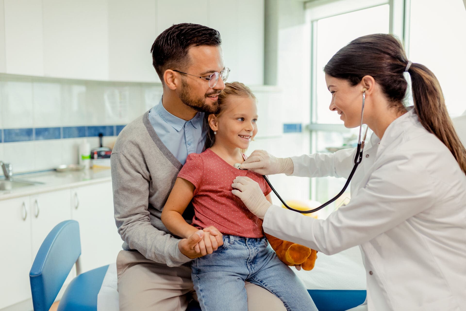 a doctor listening to a child's heart with a stethoscope while the child sits on father's lap in a pediatric urgent care setting