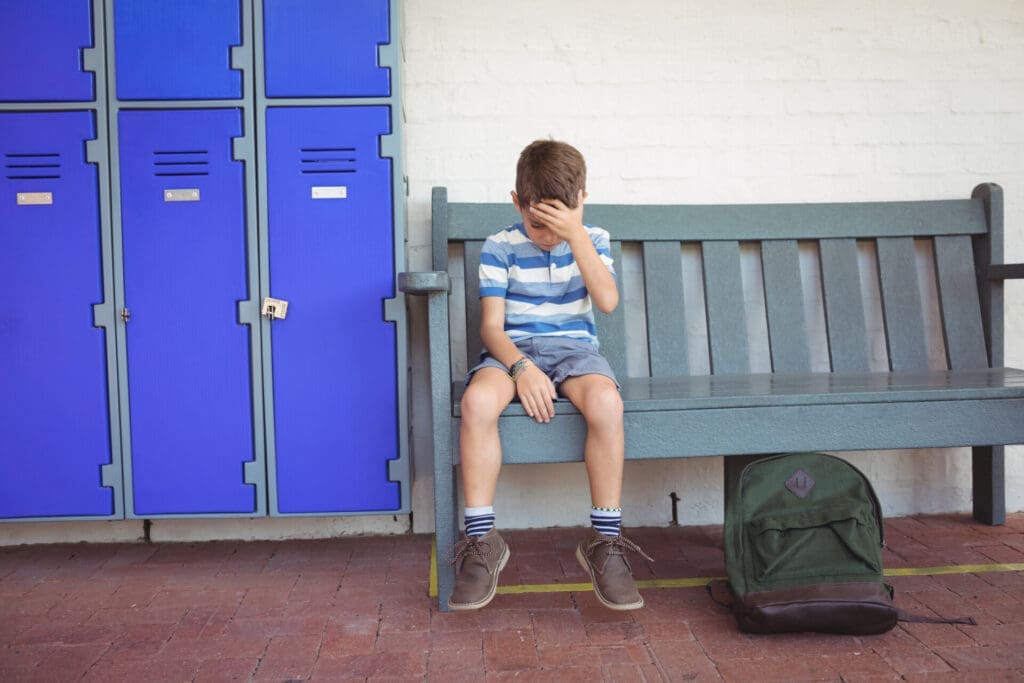 Full length of sad boy sitting on bench by lockers at school