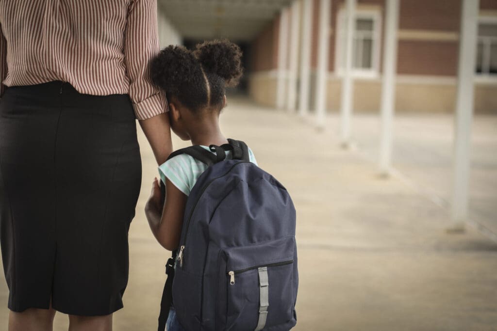 Young girl with backpack leaning on arm of adult at school.