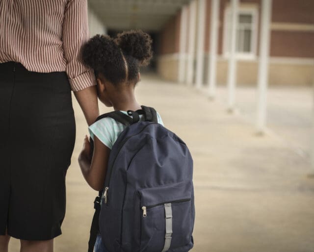 Young girl with backpack leaning on arm of adult at school.