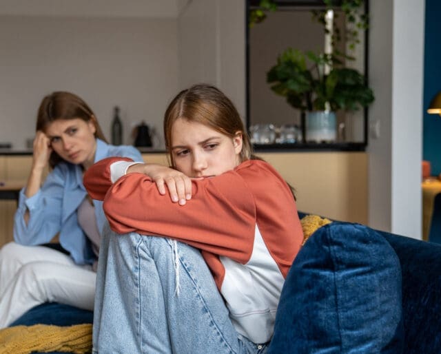 Frustrated teenage girl sits hugging his knees, looks away. Mom sits next daughter and comforts after quarrel. Communication problems between parent and kids.Ignoring children problems.Bulling school
